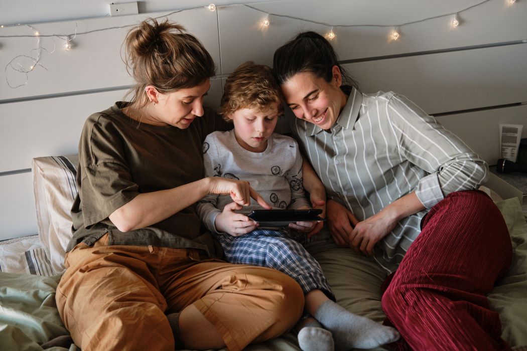 Zwei Mütter liegen mit dem Sohn auf der Couch und zeigen ihm etwas am Tablet. 